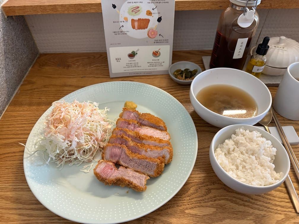 나무 탁자에 돈까스와 양배추 샐러드가 올라간 접시가 있고, 밥과 장국, 소스통과 수저 등이 놓여 있다.