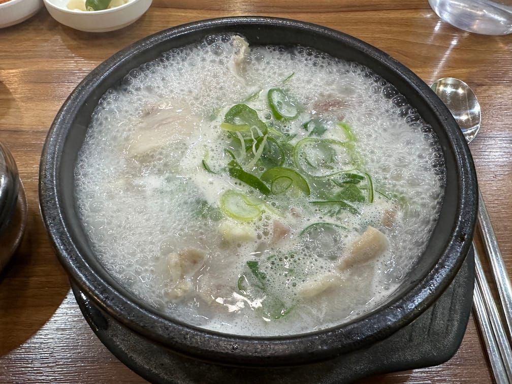 나무 테이블 위에 검은 뚝배기가 올라가 있고 그 안에 뽀얀 국물이 끓고 있는 사진. 국물 속에 채썬 파와 고기가 보인다.