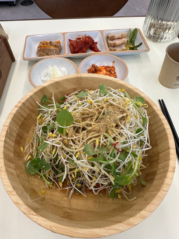 테이블에 놓인 나무 그릇 안에 담긴 면과 숙주나물, 어린 잎채소, 그 위의 들깨가루. 그 외에 김치, 절인 무, 수육, 코다리, 메밀전병 반찬이 놓여 있는 사진.
