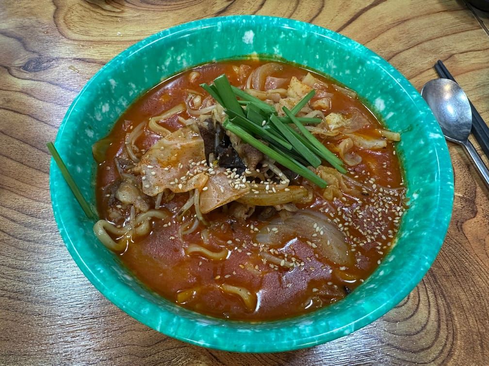 나무 테이블 위에 차려진 녹색 분식그릇 안에 담긴 붉은 국물과 양파, 고기, 굵기가 일정하지 않은 면, 목이버섯 위에 뿌려진 ㅂ