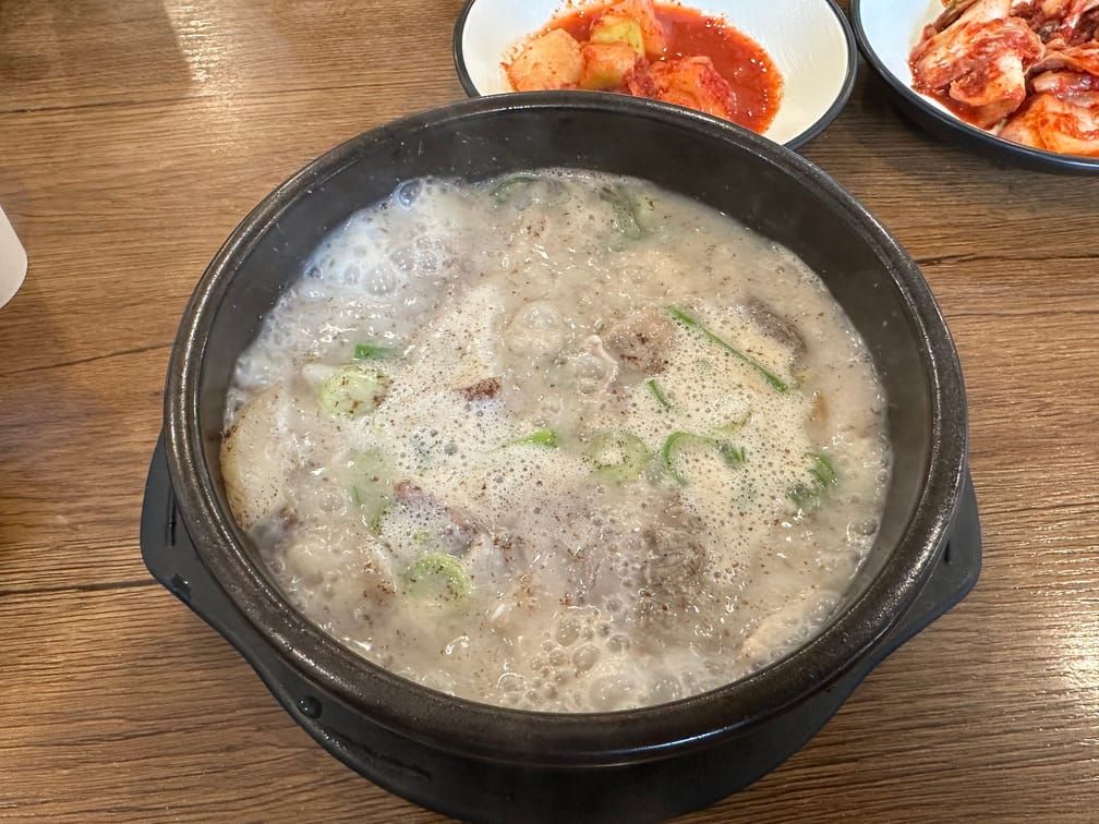 나무 테이블 위에 올라온 뚝배기 안의 뽀얀 국물과 끓고 있는 국물, 채썬 파, 후추가루. 깍두기와 김치가 담긴 그릇도 보인다.