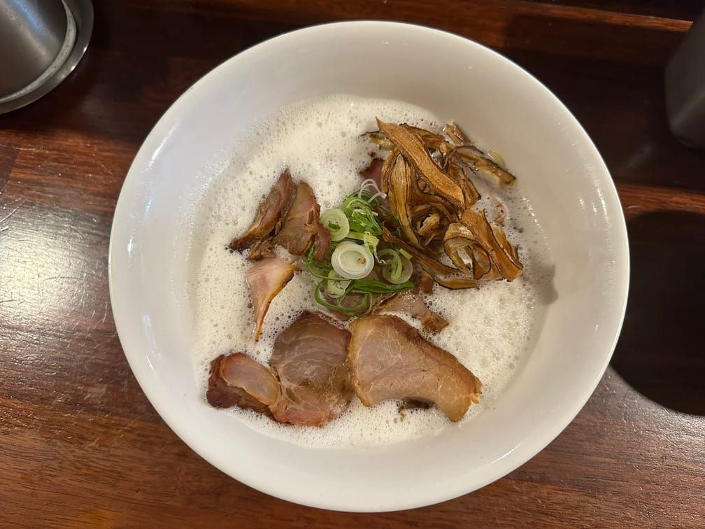 식탁에 올라온 하얀 면기 위에 하얀 거품이 가득 차 있고, 그 위로 익힌 돼지고기, 익힌 멘마, 채썬 파가 올라와 있는 사진.