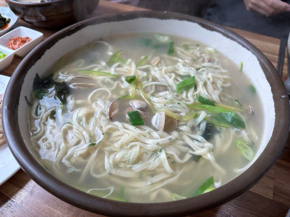그릇에 담긴 흰 뿌연 국물에 칼국수, 조개, 파, 다시마, 애호박 등이 떠 있는 모습.