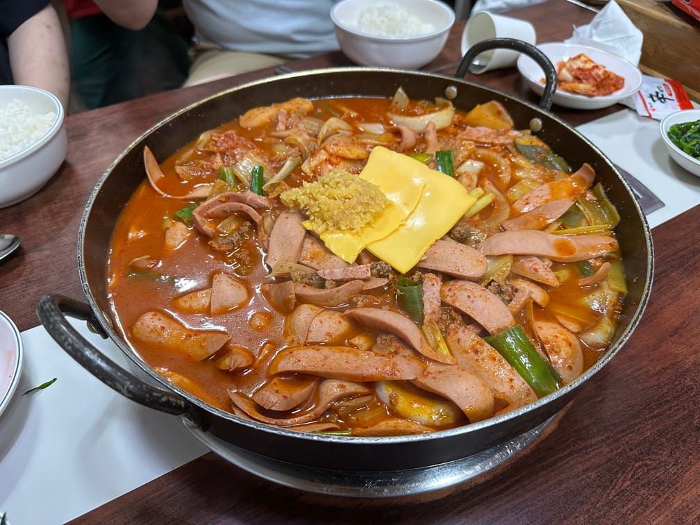 나무 식탁의 화구에 찌개 그릇이 올라와 있고 그 안에 붉은 국물, 소세지, 치즈, 다진마늘, 썬 대파등의 재료가 들어가 있다.