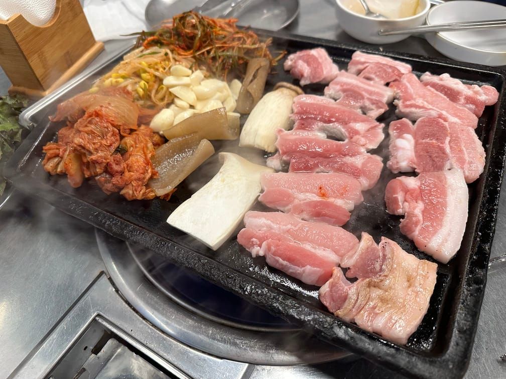 철판 위에서 익고 있는 삼겹살, 버섯, 돼지껍데기, 마늘, 콩나물무침, 배추김치, 파김치.