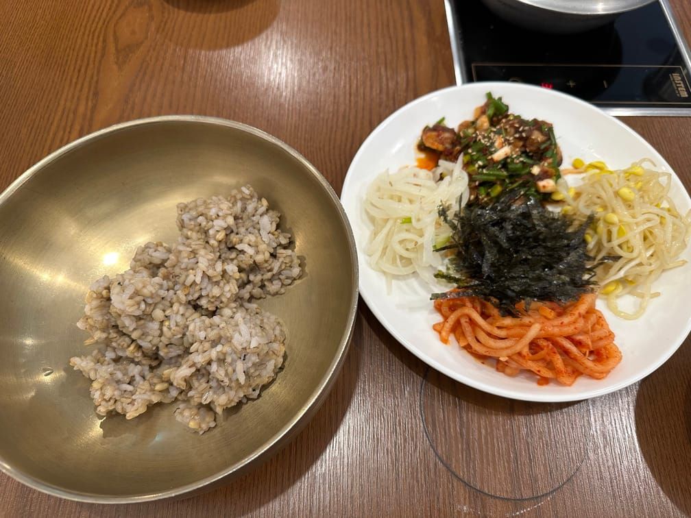 식탁 위에 유기색 그릇에 담겨있는 보리밥과 흰 접시에 차려진 김가루, 무말랭이, 콩나물, 꼬막무침 등.