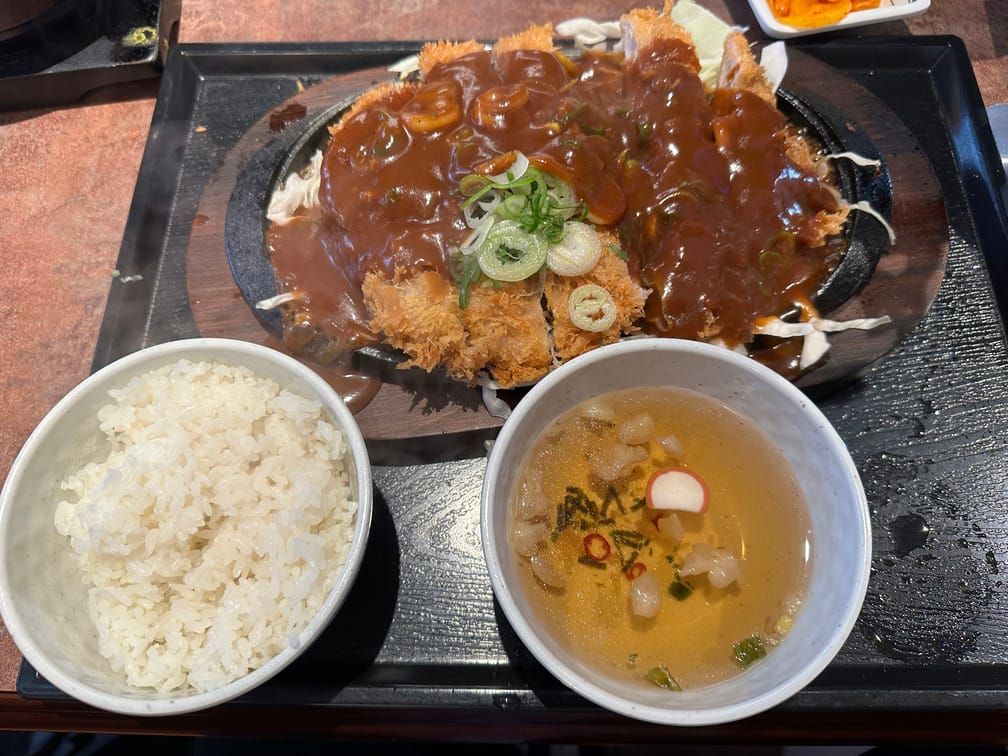 사각 쟁반 위의 흰밥, 맑은 국물, 철판 위의 돈가스와 그 밑의 양배추, 위에 끼얹어진 소스.