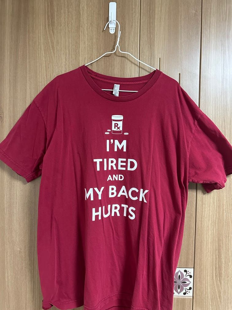 붉은 색 티셔츠에 흰 색으로 약통과 알약, I'M, TIRED, AND, MY BACK, HURTS라고 위에서 아래로 적혀있다.
