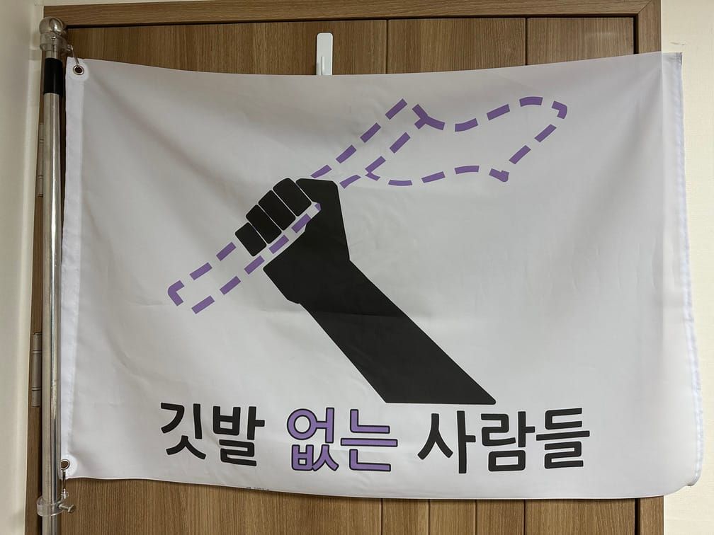 깃발 없는 사람들이라는 문구와, 깃발을 든 팔이 그려진 도안의 깃발이 벽에 걸려 있다. 깃발을 든 팔은 검은색으로, 깃발은 색 없이 연보라색 점선 외곽선으로 그려