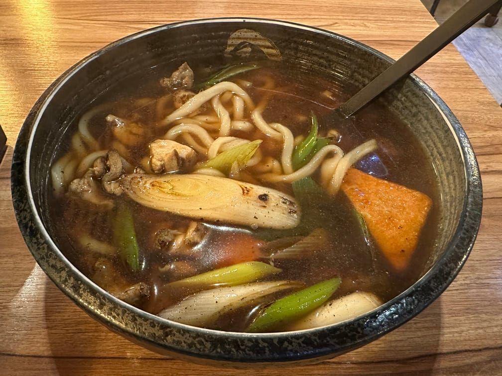 넓은 면기에 우동이 차려져 있다. 국물은 진하고, 유부, 구운 대파, 닭고기 등이 어지럽게 들어가 있다.