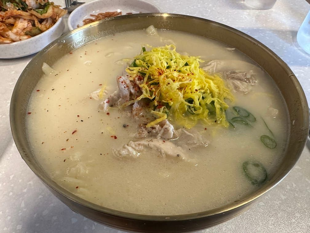 유기색 그릇의 연한 노란 국물 안에 면과 닭고기가 잠겨 있고 채썬 대파, 지단, 고추가루 등이 떠 있다.