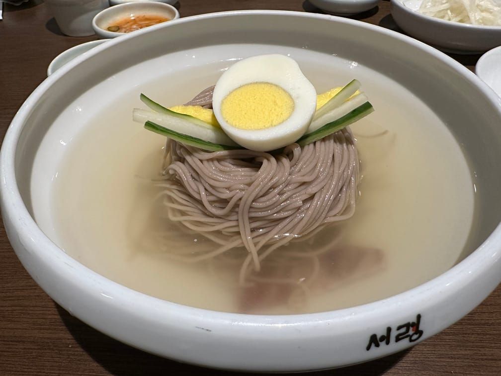 "서령"이라는 점포명이 인쇄된 그릇에 담겨 있는 맑은 국물의 메밀면. 위에는 계란 반쪽, 오이, 무, 계란 지단이 올라와 있고 바닥에는 고기가 잠겨 있다.