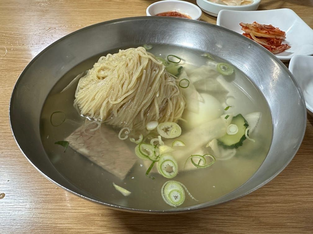 금속 면기에 담긴 냉면. 무채, 편육, 썬 파, 썬 오이가 맑은 국물에 들어가 있고 메밀면 타래가 반쯤 담겨 있다.