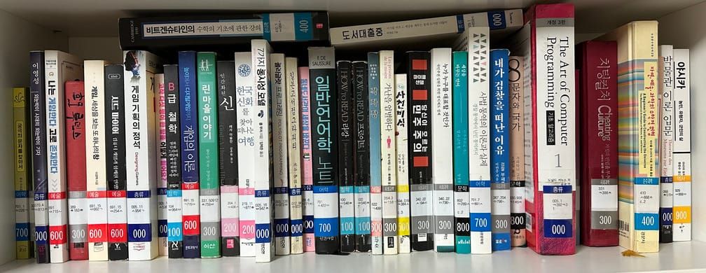 흰 책장에 도서관의 분류표가 붙어있는 책이 40권 가까이 빼곡히 꽂혀있는 모습.