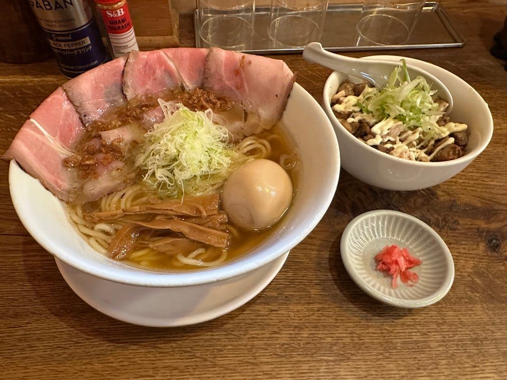 나무 식탁에 챠슈와 달걀이 올라간 반투명한 국물의 라멘과 마요네즈가 뿌려진 고기가 보이는 밥공기, 초생강이 차려진 모습의 사진.