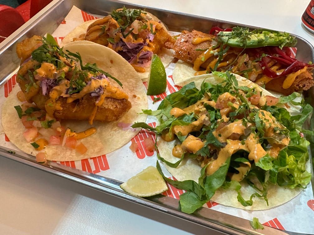 식탁 위의 은색 트레이 위에 놓인 4개의 타코. 또띠야 위에 야채, 살사 소스, 여러 재료, 라임 조각이 놓여 있다.