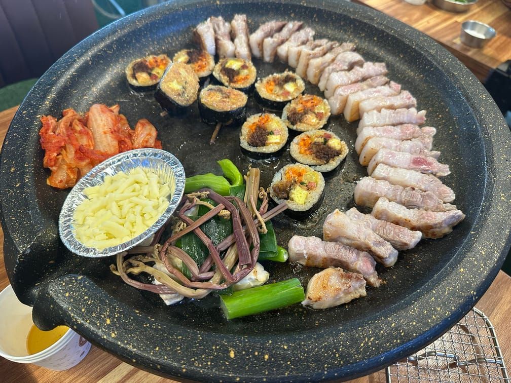 둥그런 구이판에 삼겹살, 김밥, 고사리 등의 야채, 김치, 치즈 등이 차려진 모습.