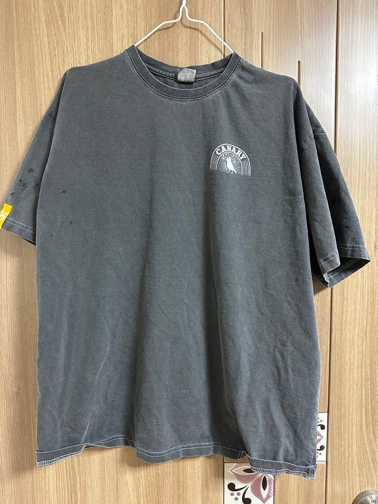 회색 거친 재질에 헬멧을 쓴 카나리아와 반원형으로 'CANARY PROJECT'라는 로고가 왼쪽 가슴에 적힌 티셔츠.