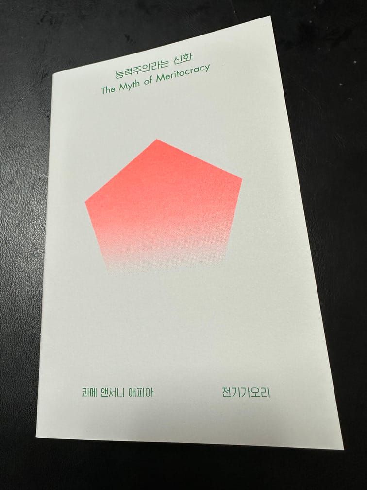 검은 배경 위의 흰 표지의 책 『능력주의라는 신화』. 아래쪽이 희미해지는 분홍색 정오각형이 가운데에 있는 심플한 디자인.