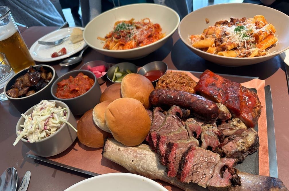 테이블에 고기 플래터, 파스타, 맥주잔, 개인 접시 등이 올라와 있는 사진.