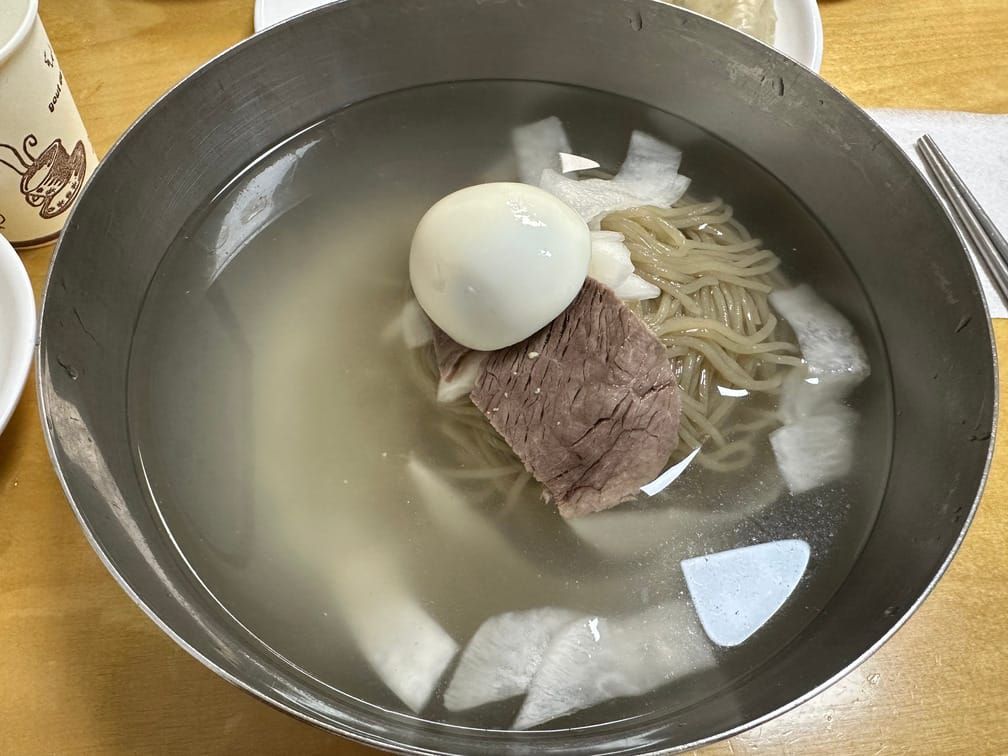 식탁 위에 올라와있는 금속 면기에 반투명한 맑은 국물, 무절임, 메밀면, 편육, 달걀 반쪽 구성의 냉면이 담겨 있다.