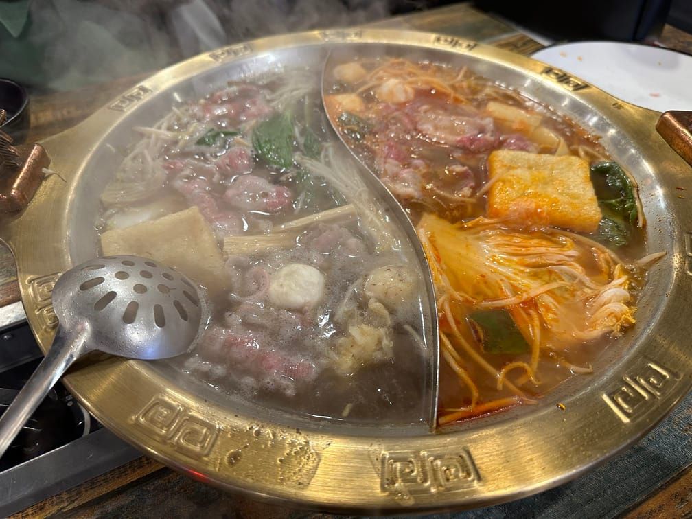문양이 새겨진 훠궈 냄비의 반으로 갈린 칸에서 왼쪽에는 맑은 국물, 오른쪽에는 붉은 국물 위에 고기, 배추, 버섯, 완자, 유부 등이 떠 있는 사진.