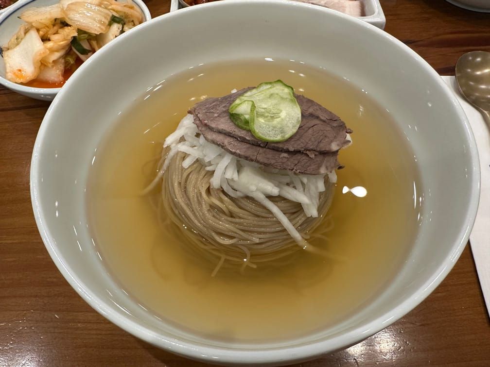 테이블 위의 흰 면기에 담긴 투명한 노란 국물, 면 뭉치, 채썬 무절임, 수육, 얇게 썬 오이 사진.