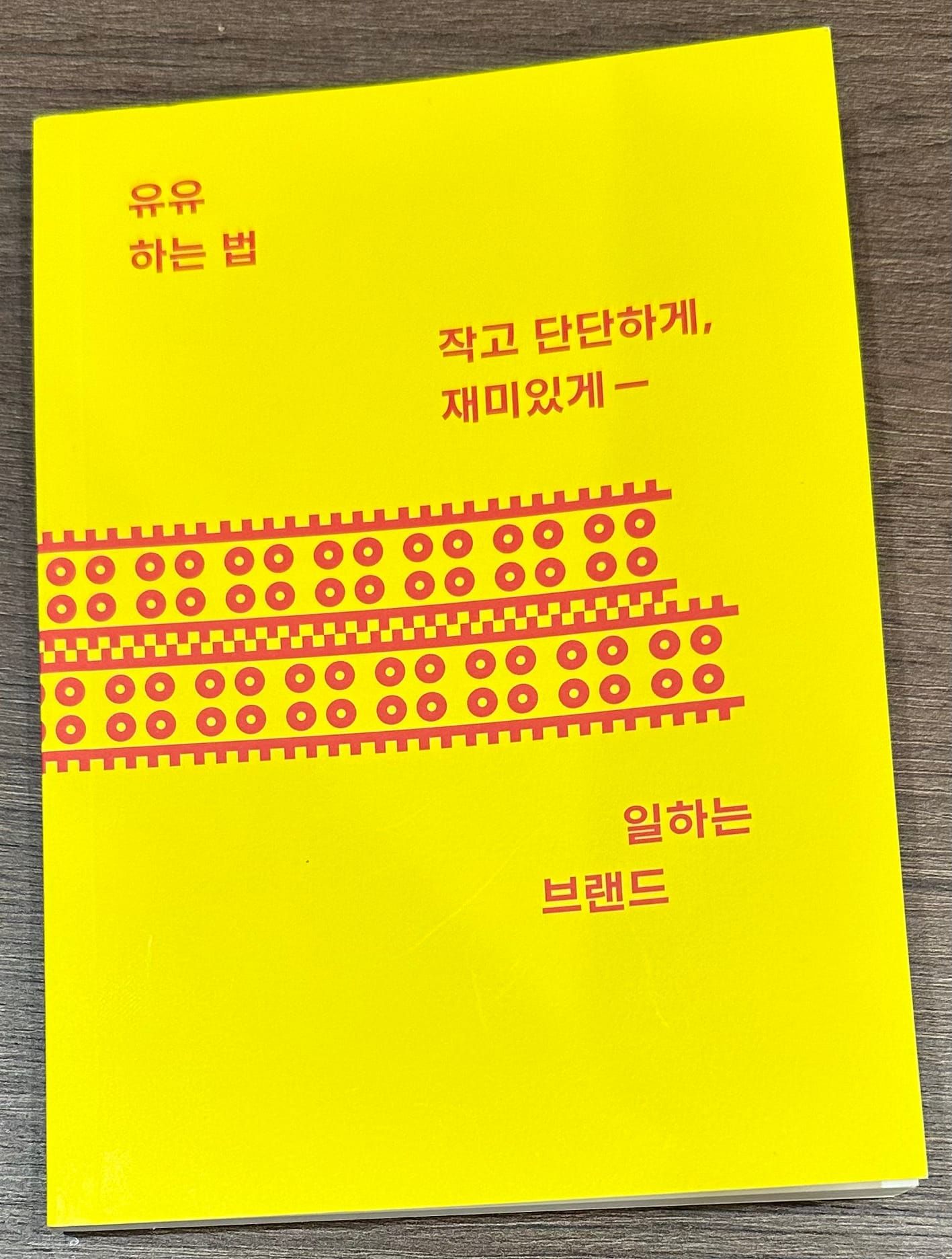 나무 책상에 놓인 책 『유유 하는 법』의 사진. 책 표지는 노란 배경의 '유유'라는 글자들이 맞물린 벽돌처럼 배치되어 있는 디자인과 책의 제목, 부제가 붉은 글씨로 구성되어 있다.