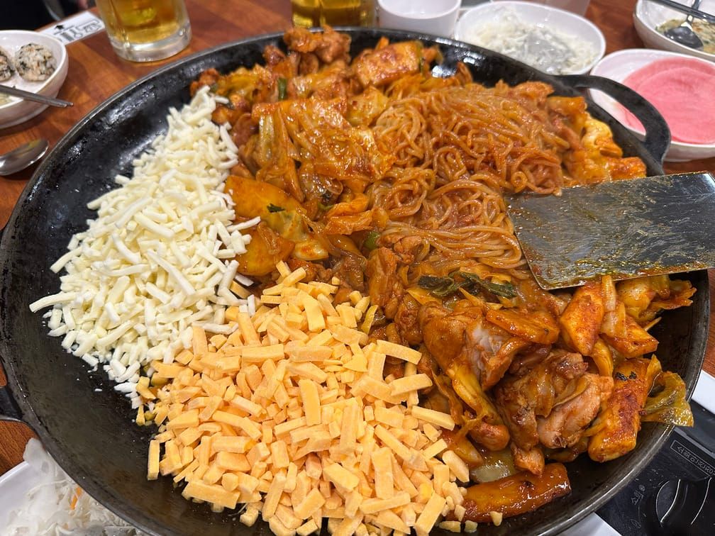 식탁에 놓인 넓은 냄비에 올라가있는 닭갈비와 고체 치즈들의 사진.