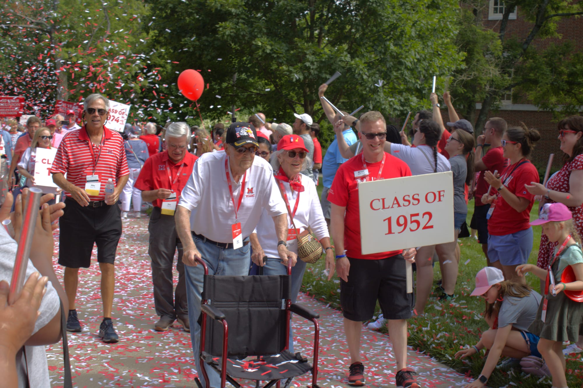 Carl Patzer walks down a sidewalk covered in red and white confetti with spectators on both sides, helped by a walker. On the right, a man holds a sign that reads "Class of 1952."