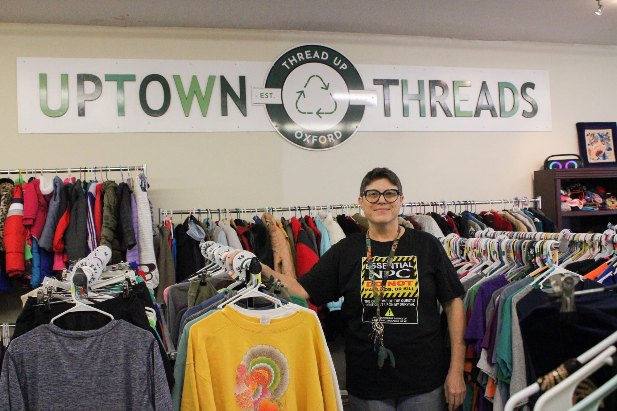 Shana Rosenberg stands surrounded by clothing racks underneath a sign that reads "Uptown Threads."