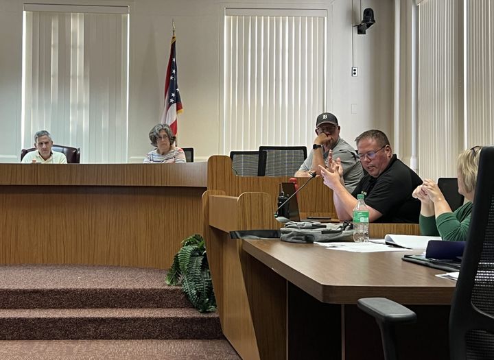 Three board members sit at a raised table. Police Chief John Jones is speaking to the right at a lower table.