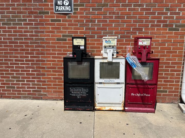 Three old and worn newsstands sit on an empty sidewalk, one labeled "The Oxford Press"