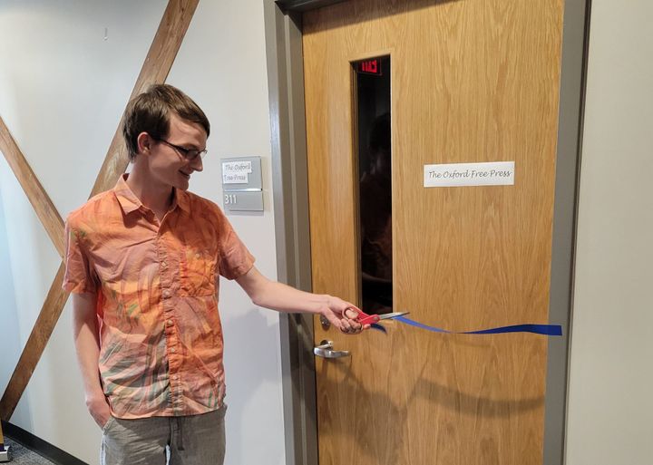 Sean Scott stands on the left of a closed door with an "The Oxford Free Press" sign and cuts a blue ribbon with scissors.