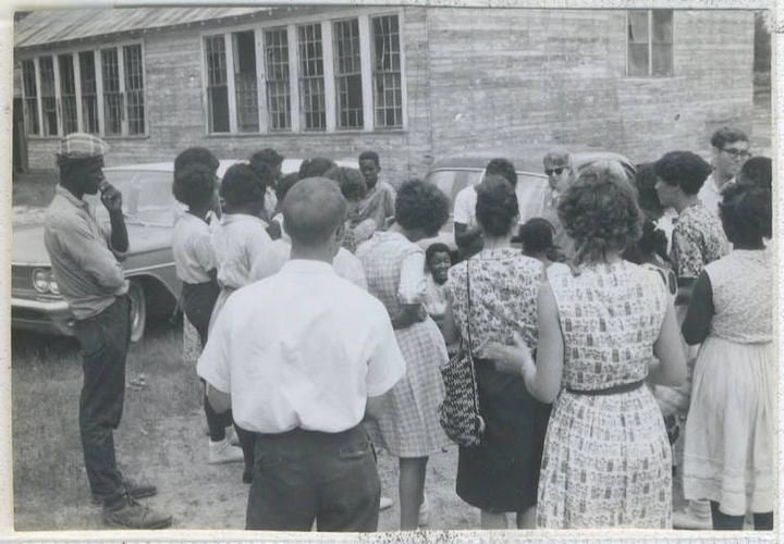 A group of people stand outside in front of several cars. The photo is black and white