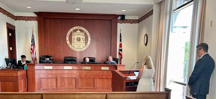 3 Commissioners sit behind their curved wooden desk, adorned with name plaques, and discuss the agenda for the day.