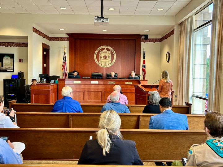 Two commissioners behind rounded desk hear woman in orange speak as well as different presenters for the day.