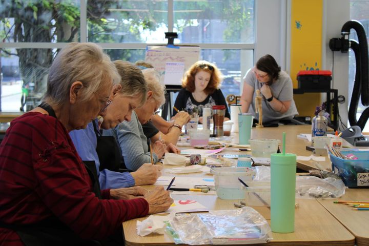 Several women paint at a cluttered table in a brightly lit room