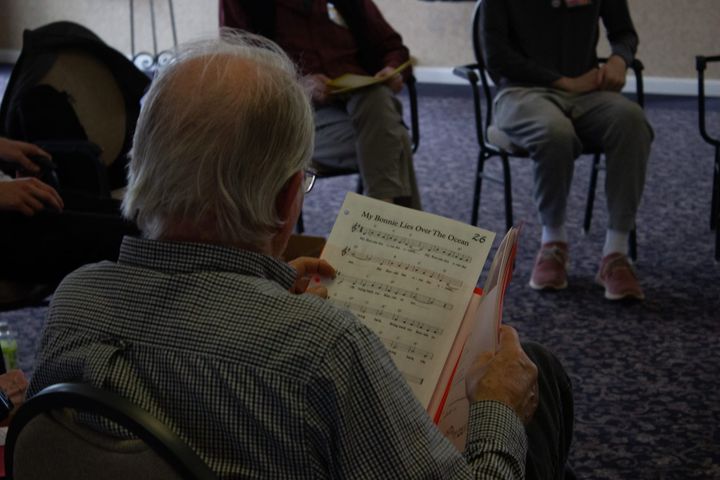 A choir member reads sheet music labeled "My Bonnie Lies Over The Ocean"