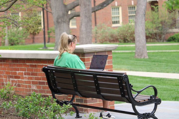 A woman in a green shirt sits on a bench working on her laptop