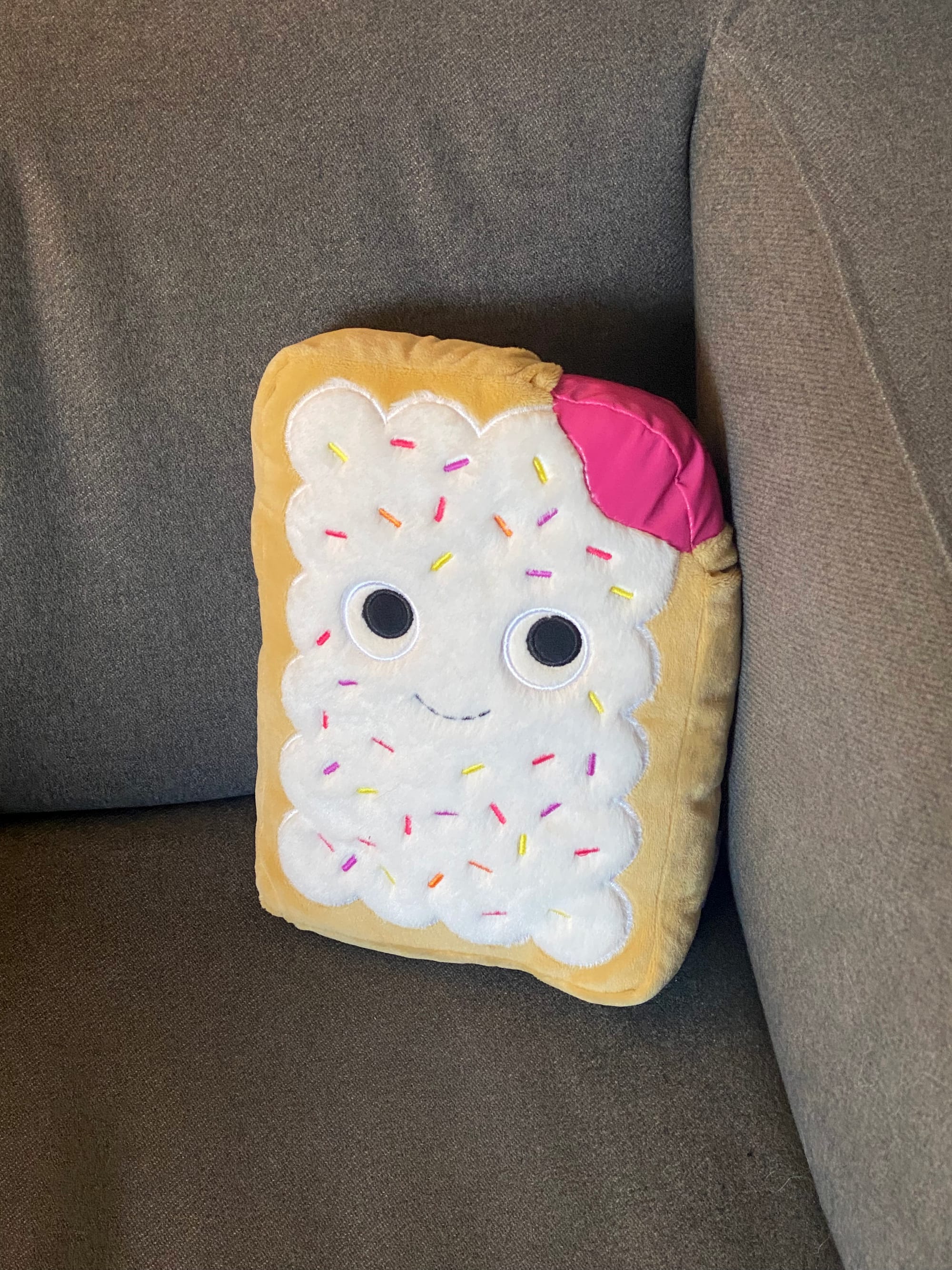 A stuffed toy pop tart with expressive eyes, resting in the corner of an grey armchair.