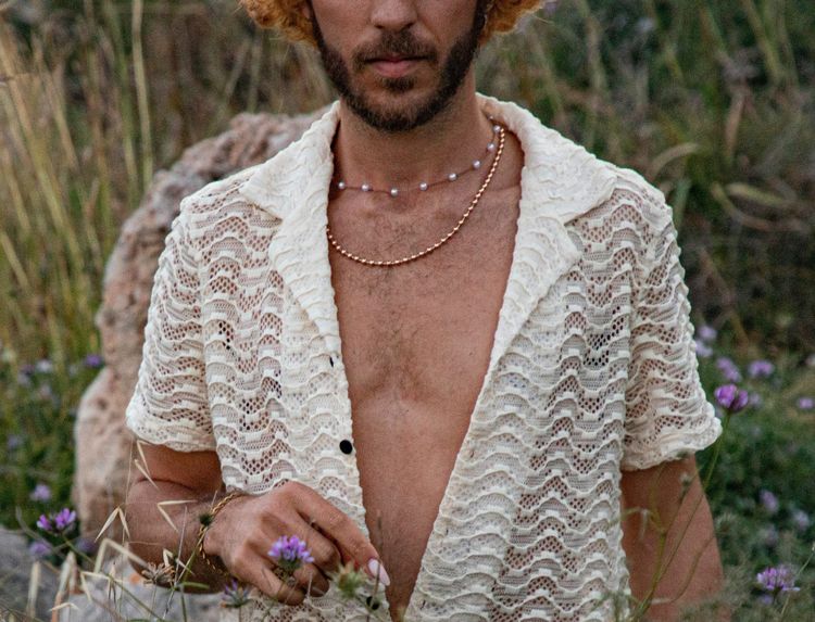 Man with a knitted short sleeve shirt open to reveal two necklaces around his neck.