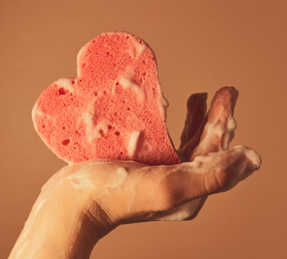 Hand holding a heart-shaped sponge, both covered in lather.