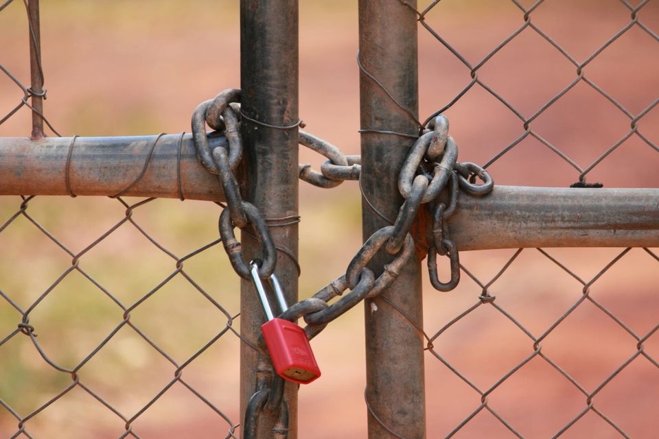 A chain in the shape of a heart, fastened by a red lock, and holding two sections of a fence together.