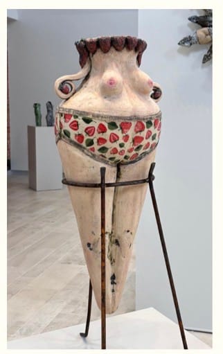 A large ceramic vase shaped like a stylized human torso, featuring painted strawberries on the midsection. It stands on a metal tripod in a gallery.