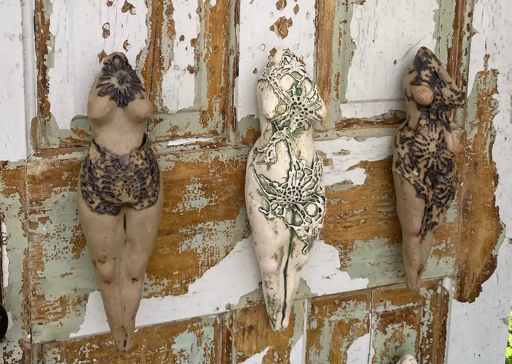Three sculpted female torsos with intricate lace-like designs hang on a weathered, peeling wooden door. The scene conveys an artistic, rustic vibe.