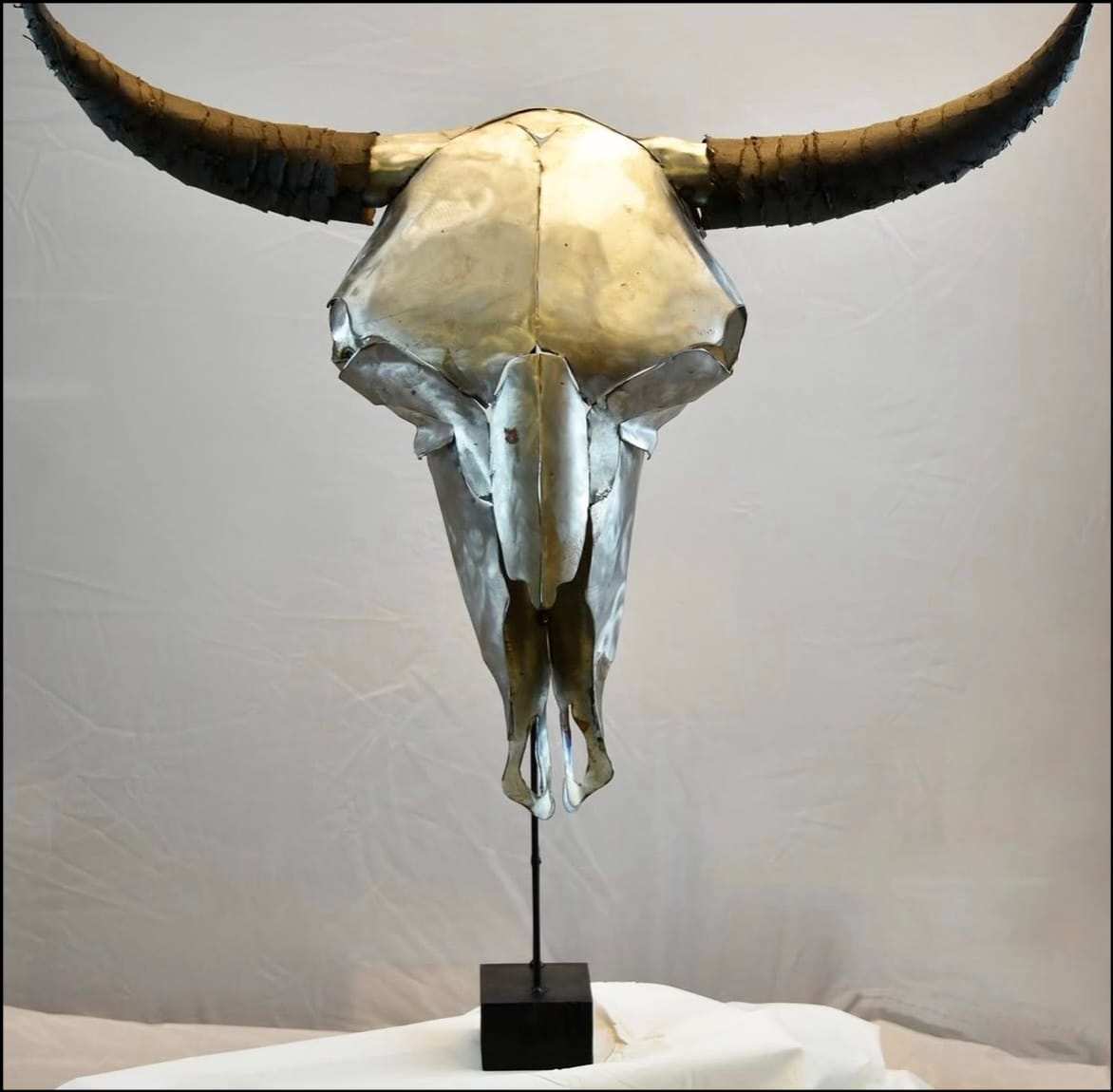 A metallic sculpture of a bull skull with large curved horns is displayed on a black stand against a plain background.