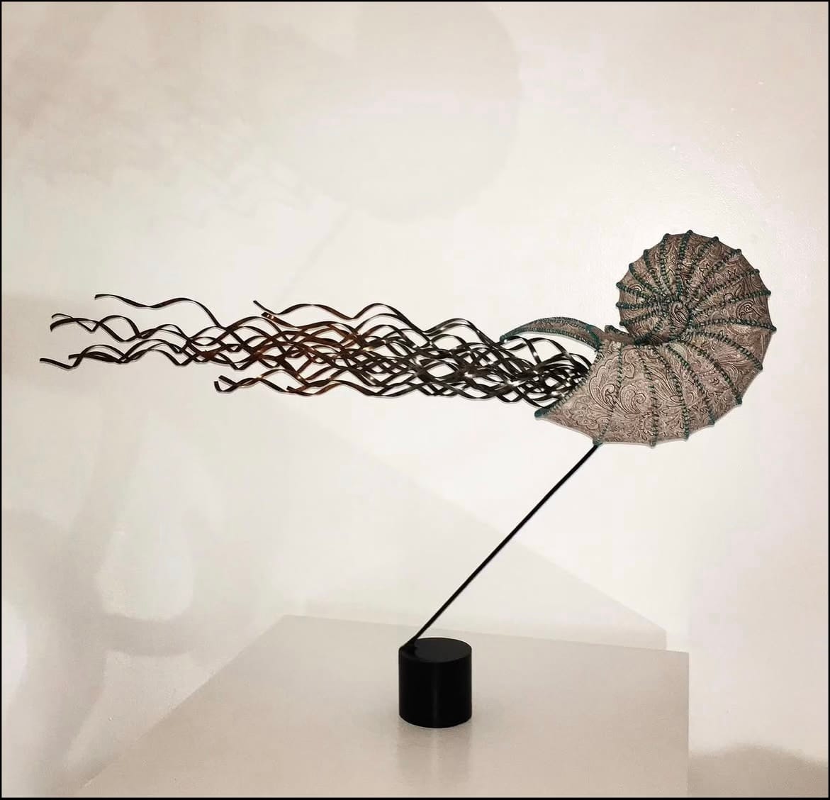 Sculpture of a Nautilus shell trailing steel streamers or tentacles. Sculpture is supported by a thin metal rod. 