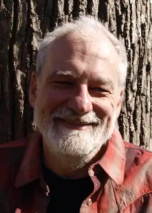 A man with white hair and a beard smiles warmly, wearing a red plaid shirt. He stands in front of a large tree trunk, in natural sunlight.
