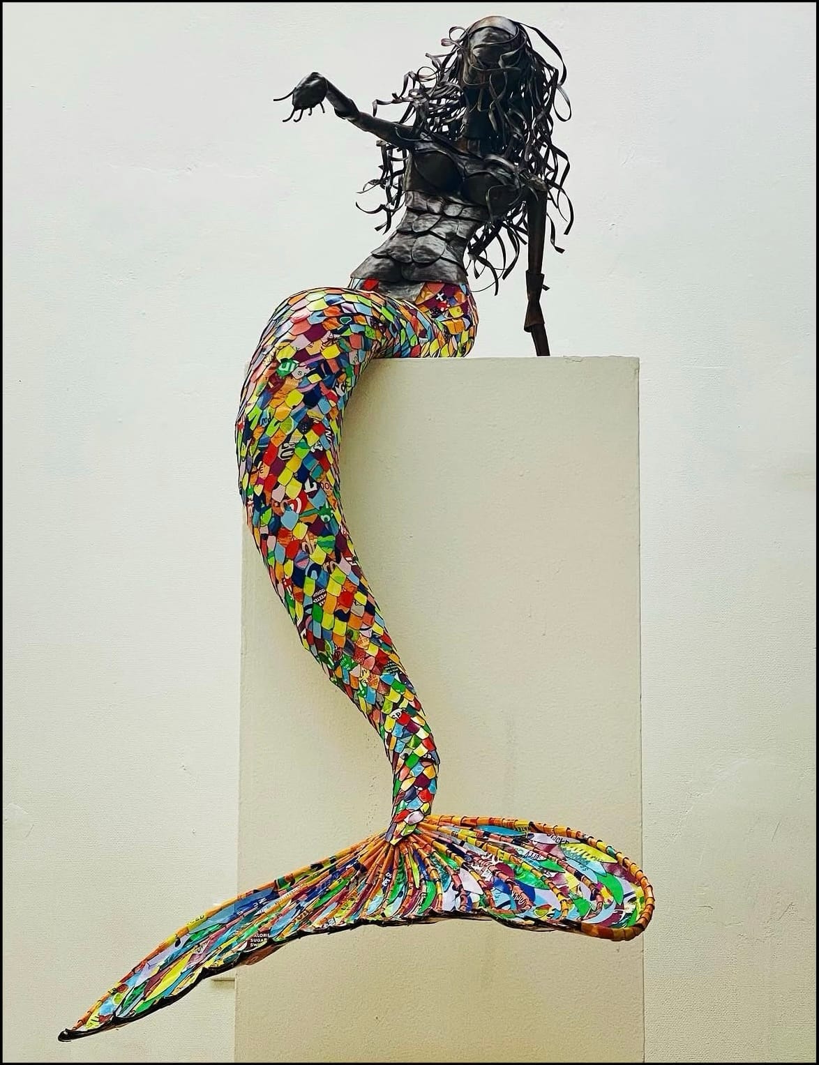 Sculpture of a mermaid with a dark metallic gray head and torso. The tail and tail-fin are a multicolored mosaic of cardboard pieces.
